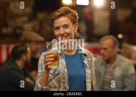 Portrait of young woman holding glass of beer and smiling at camera while standing in the pub Stock Photo