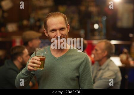Portrait of young bearded man drinking beer and smiling at camera while standing in the pub Stock Photo