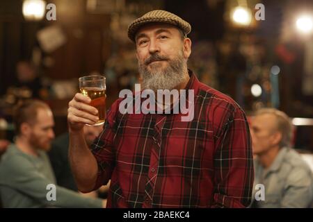 Mature bearded man in casual clothing holding glass of beer and looking at camera while resting in the pub with his friends Stock Photo