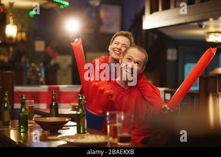 Happy young couple of fans in red shirts watching football match and cheering for their team in sport bar Stock Photo