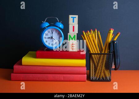 A stack of books, a green flower, blue clock and a glass of pencils, and wood cubes with text time stand on an orange background Stock Photo
