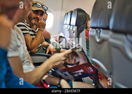 Young man with a short beard wearing a cap, sunglasses and bracelet on an airplane looking at the magazine that another traveler shows him. Horizontal Stock Photo