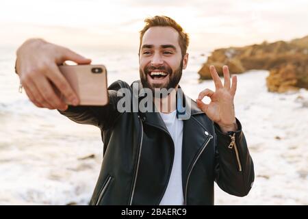 Image of young handsome man in leather jacket taking selfie photo on smartphone while walking by seaside Stock Photo