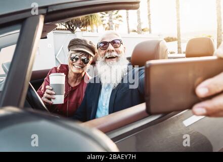 Happy senior couple taking selfie on new modern convertible car - Mature people having fun together making self photos during road trip vacation Stock Photo