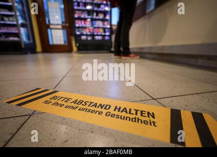 27 March 2020, Saxony, Leipzig: A woman is standing behind a warning sticker asking for distance at the checkout in a supermarket. Grocery stores are open despite contact restrictions. Protective walls and safety notices are to protect employees and customers from the corona virus. Photo: Jan Woitas/dpa-Zentralbild/dpa Stock Photo