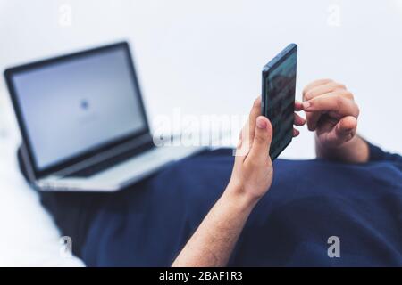 close up view of a man using his mobile phone and a lap top while lying on bed Stock Photo