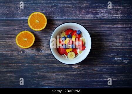 Breakfast cereal bowl with fresh fruits (strawberry, banana, blueberry, mango). Half-cut orange for decoration. Dark wooden table background. Stock Photo