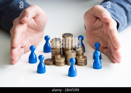 Businessperson's Hand Protecting Blue Human Figures Surrounding Stacked Golden Coins Over Desk