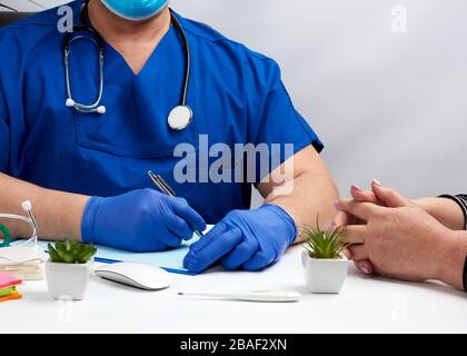 male doctor in uniform and in blue latex medical gloves sits at the table and carries out the reception of patients, workplace and office of the docto Stock Photo