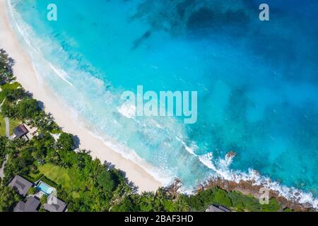 Aerial top-down drone view of turquoise waves on white sand beach with resort and tropical trees at Anse Petite, Mahe, Seychelles Stock Photo