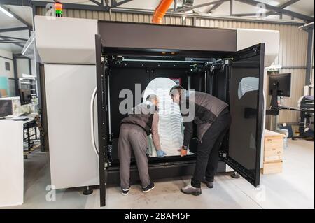 Two technicians wear a huge 3d model of the sneaker printed on a large modern 3d printer machine with computer control Stock Photo
