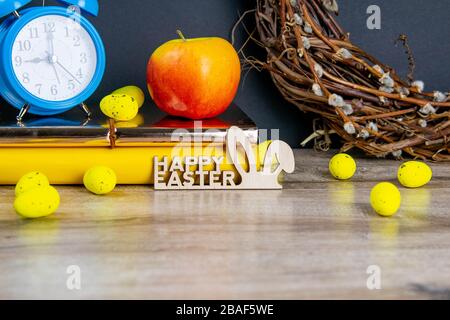 Easter concept, clock and apple stand on yellow books, eggs are scattered next to them, there is a wreath in the background Stock Photo