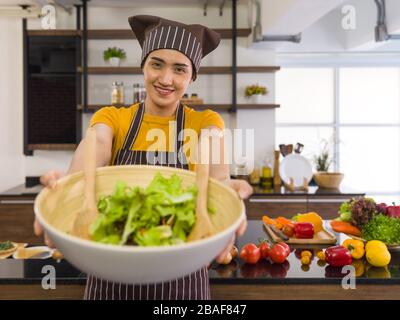 The housewife dressed in an apron and a hair cap, smiled and offered a salad bowl on the front. Morning atmosphere in a modern kitchen. The kitchen co Stock Photo