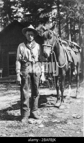 Cowboy in America with his saddled horse. He is unarmed. The log cabin behind him has a sign that seems to say 'Barack     '  This might be a barracks building for cowboys or a camp or ??  An indicator that he might not be a real cowboy is that he is wearing shoes, not boots.   To see my Western-related  images, Search:  Prestor  vintage  west  vehicle Stock Photo