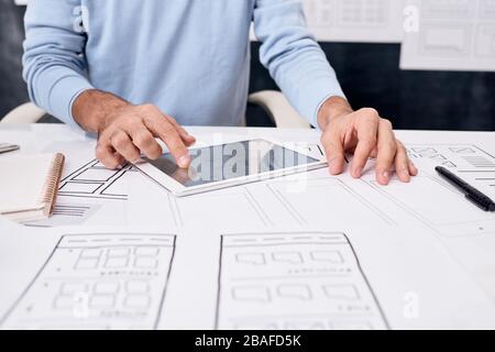 Close-up of unrecognizable man sitting at table with sketches and using tablet while developing app design Stock Photo