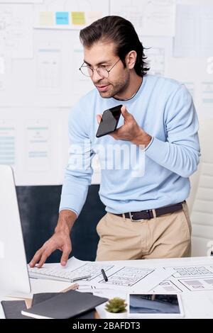 Modern middle-aged man with stubble standing at computer and using smartphone while recording voice message for colleague Stock Photo