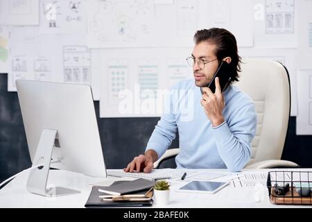Content brown-haired manager of IT company sitting at desk and talking to colleague by phone while analyzing data on computer Stock Photo