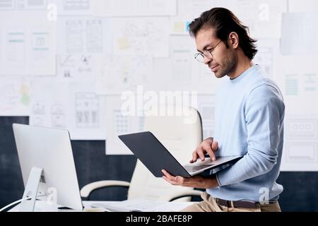 Serious modern middle-aged man in glasses using laptop while analyzing interfaces of web sites in UI design office Stock Photo
