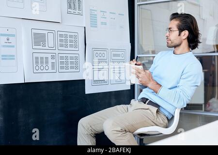 Pensive creative designer in glasses looking at board with sketches and making notes about user interface design Stock Photo