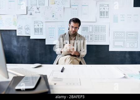 Busy fashionable man in glasses sitting against board with interface sketches and working on new design project Stock Photo