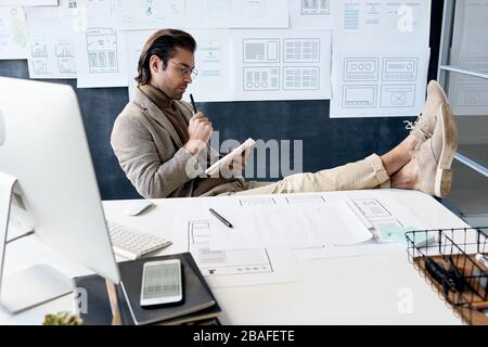 Pensive handsome man in glasses focused on tasks sitting with feet on table and analyzing own notes Stock Photo