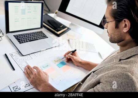 Serious phone interface engineer in glasses sitting at desk and making notes on interface sketch Stock Photo
