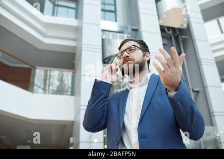 Young bearded businessman in formalwear and eyeglasses explaining something to colleague while calling by smartphone Stock Photo