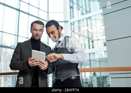 One of two young businessmen pointing at online document in tablet while explaining terms of electronic contract to colleague Stock Photo