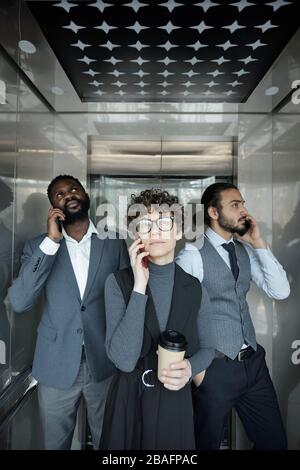 Group of three young intercultural office managers with smartphones calling clients inside elevator during coffee break Stock Photo