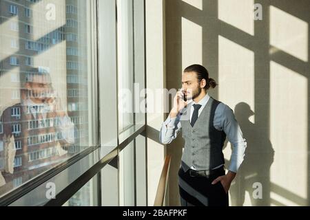 Young serious businessman looking through window on sunny day while consulting one of clients by smartphone on working day Stock Photo
