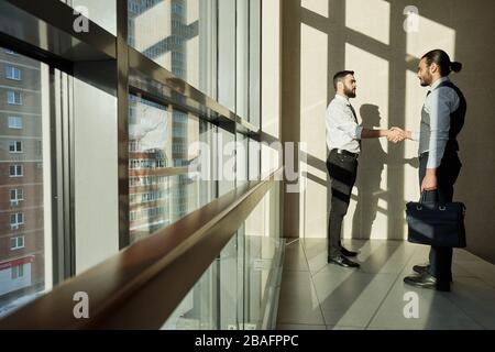 Two successful business partners shaking hands of one another while while standing by large window in office center in the morning Stock Photo