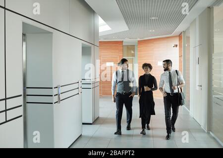Three young cheerful colleagues in formalwear chatting on their way to office while passing by elevator after coffee break Stock Photo