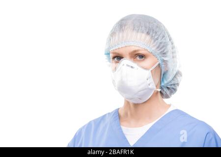 Contemporary young female doctor or nurse in protective mask and uniform standing in front of camera in isolation and looking at you Stock Photo