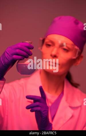 Gloved hands of young female pharmacist in whitecoat and protective eyeglasses holding beak with blue liquid substance in lab Stock Photo