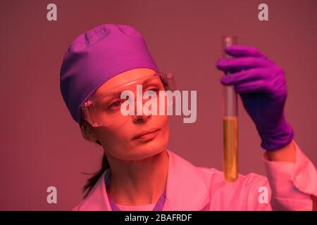 Young female laboratory worker in whitecoat, protective gloves and eyeglasses looking at yellow liquid substance in flask Stock Photo