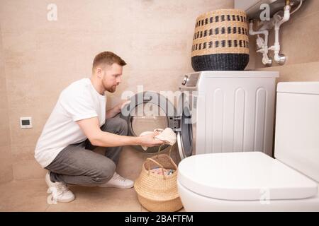 Young man taking dirty towel from basket and putting them into washing machine while squatting against wall in bathroom Stock Photo