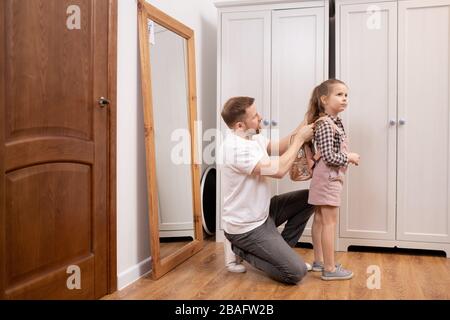 Young careful man putting small rucksack with books and copybooks on back of his cute little daughter going to elementary school Stock Photo