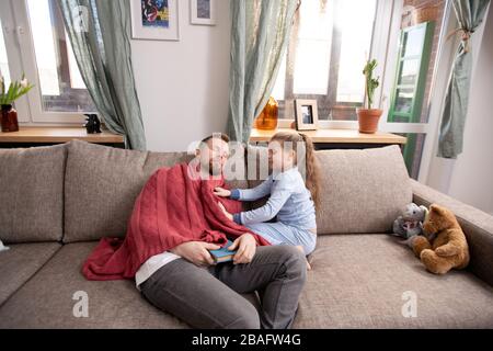 Careful cute little daughter putting warm woolen sweater on her father napping on couch after reading book of stories Stock Photo