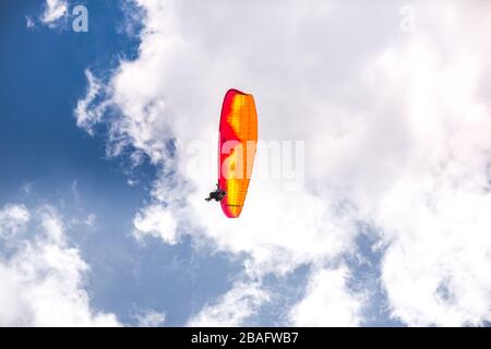 Paragliding in a bright orange and red colorful glider aircraft on a blue cloudy day Stock Photo