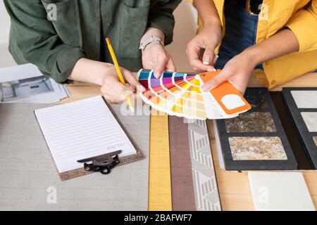 Hands of two young female interior designers consulting about choice of color for one of rooms while holding palette over table Stock Photo