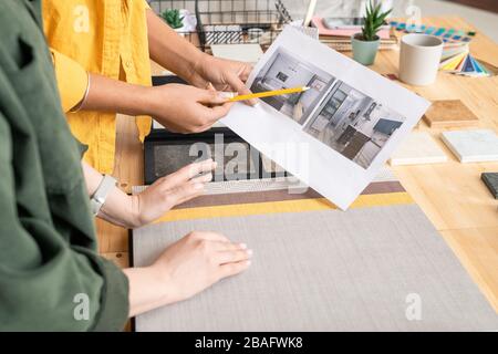 One of two young creative female designers pointing at photo of room interior on paper while discussing it with colleague in studio Stock Photo