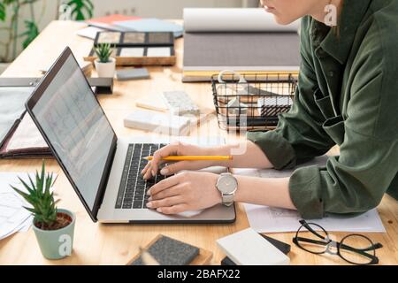 Young creative female designer bending over desk in front of laptop while working with electronic sketch of house or flat by workplace Stock Photo