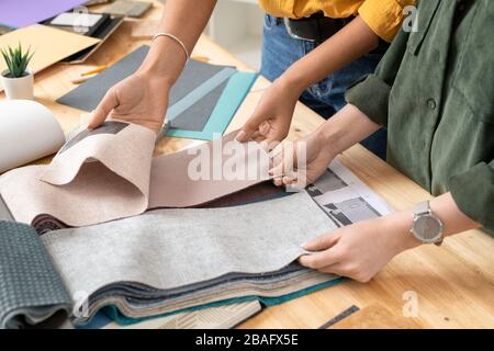 Hands of two young female colleagues looking through fabric samples by desk while choosing suitable ones for modern furniture Stock Photo
