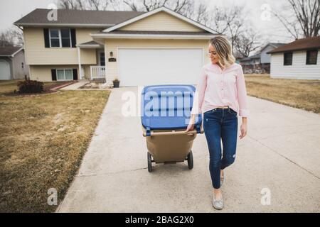 Young woman pulling recycling bin down driveway Stock Photo