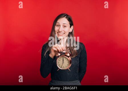 It's time. Cheerful woman is holding an alarm clock and smiling at the camera on red background. Stock Photo