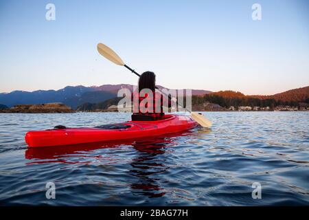 Adventurous Girl Paddling on a Bright Red Kayak in calm ocean water during a vibrant and colorful sunset. Taken in Indian Arm, Deep Cove, North Vancou Stock Photo
