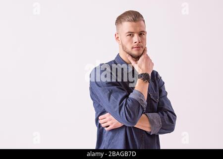 Portrait of young thoughtful man holding hand on chin and looking to camera while isolated over white background. Stylish handsome bearded hipster in Stock Photo