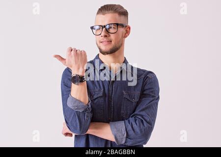 Studio shot of young male student in eyewear showing to copy space and smiling while standing against white background. Handsome bearded hipster man p Stock Photo
