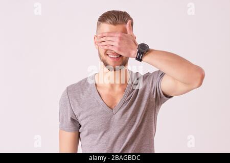 Studio shot of cheerful young man covering his eyes with hand and smiling while isolated on white background. Stylish bearded hipster standing blindfo Stock Photo
