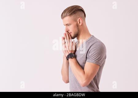 Side view of young thoughtful man holding hands clasped and looking down while isolated on white background. Bearded hipster man in t-shirt is praying Stock Photo